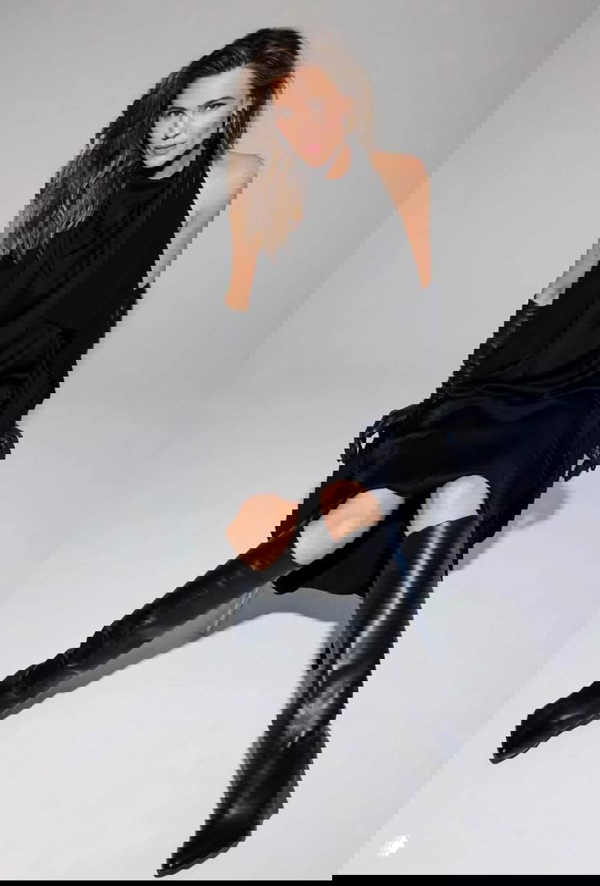 Женщина в черном платье и длинных кожаных сапогах, сидящая в студии, элегантный образ с перчатками и строгой позой.