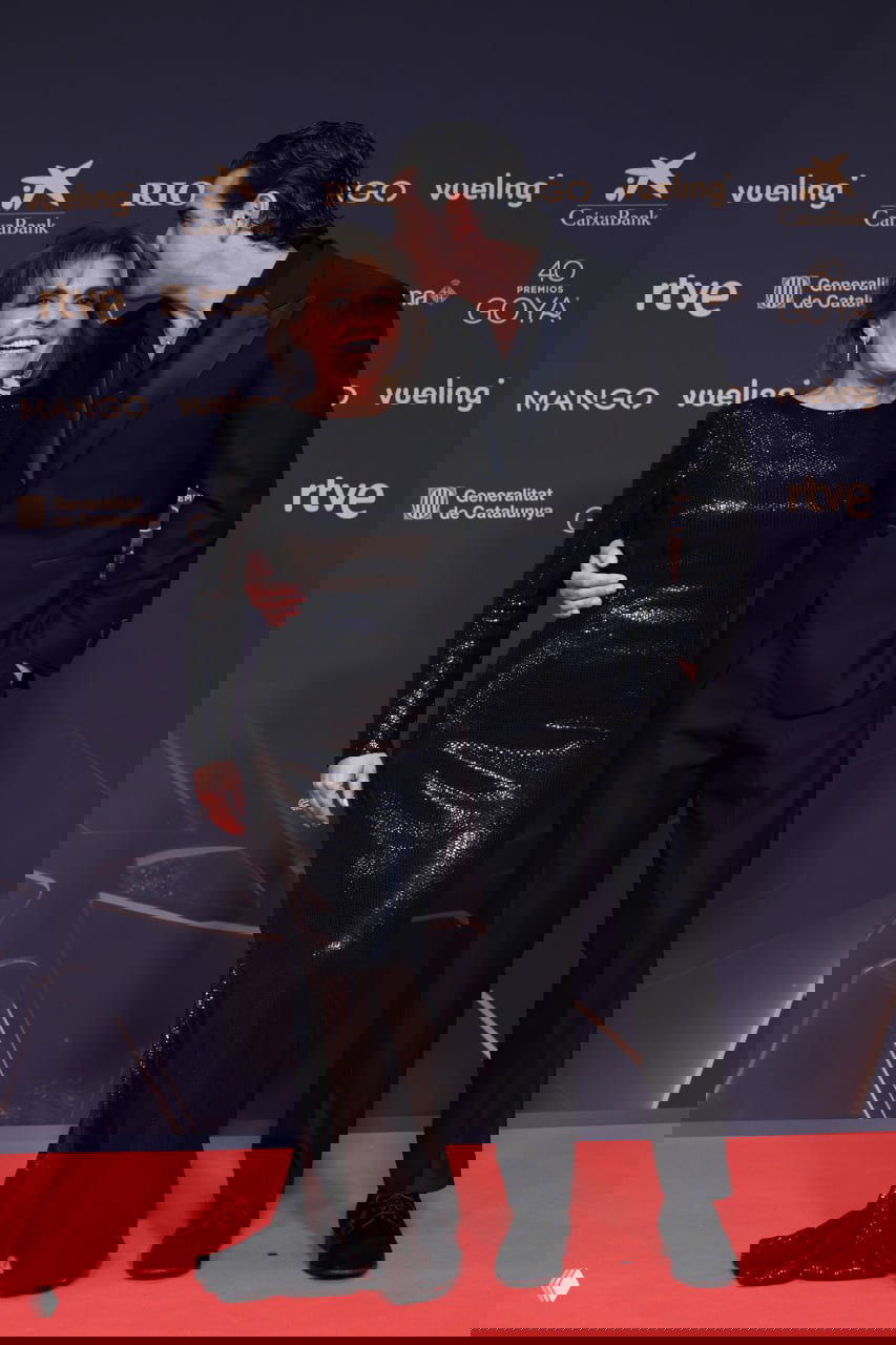 Мужчина целует женщину в чёрном блестящем платье на красной дорожке Goya в Барселоне; улыбки и фон с логотипами.