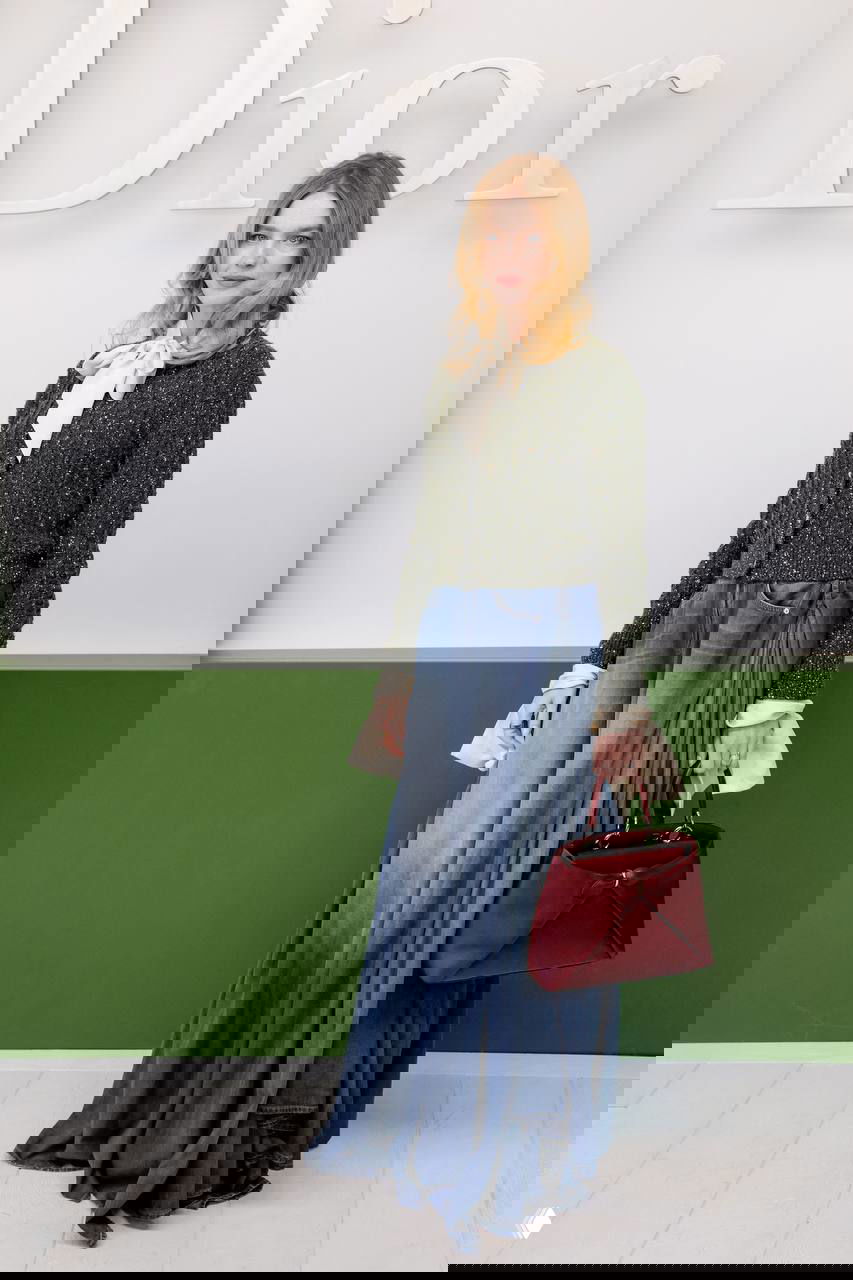 Гостья в твидовом кардигане и длинной джинсовой юбке с красной сумкой стоит на фоне зелено‑белой инсталляции и логотипа бренда.