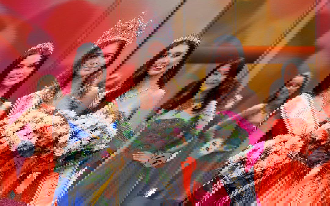 Победительницы и призёры на сцене в коронах и с букетами; видно праздничную церемонию награждения и ленты с годом конкурса.