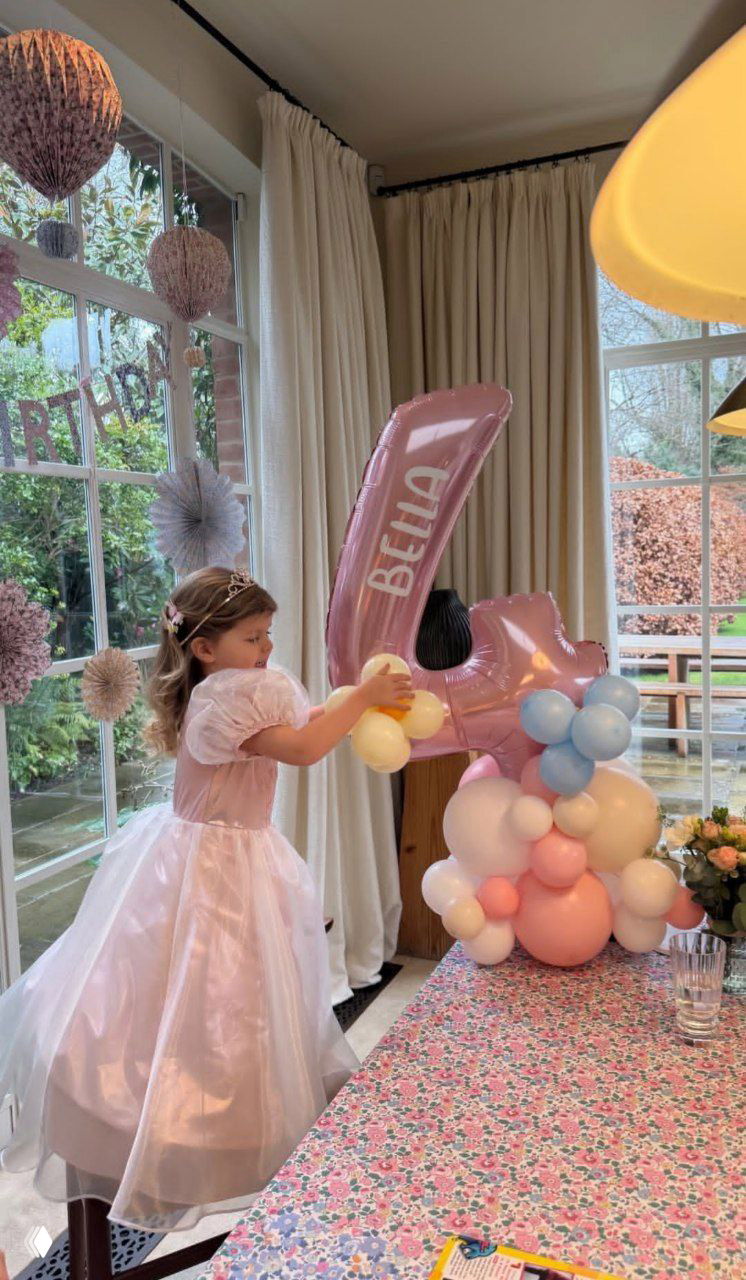 Девочка в нарядном розовом платье у стола с композициями из шаров; большая розовая цифра 4 с надписью «BELLA» видна рядом и украшает праздник.