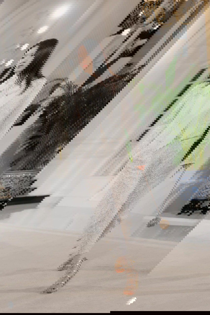 Кружевной брючный костюм молочного цвета с подъюбником и бантом на шее — элегантный образ модели на показе Chanel в светлом интерьере отеля.