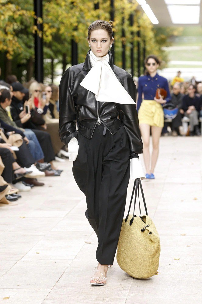 Показ на открытом воздухе: модель в чёрной кожаной куртке и белом шарфе несёт большую плетёную сумку — тренд 90‑х.