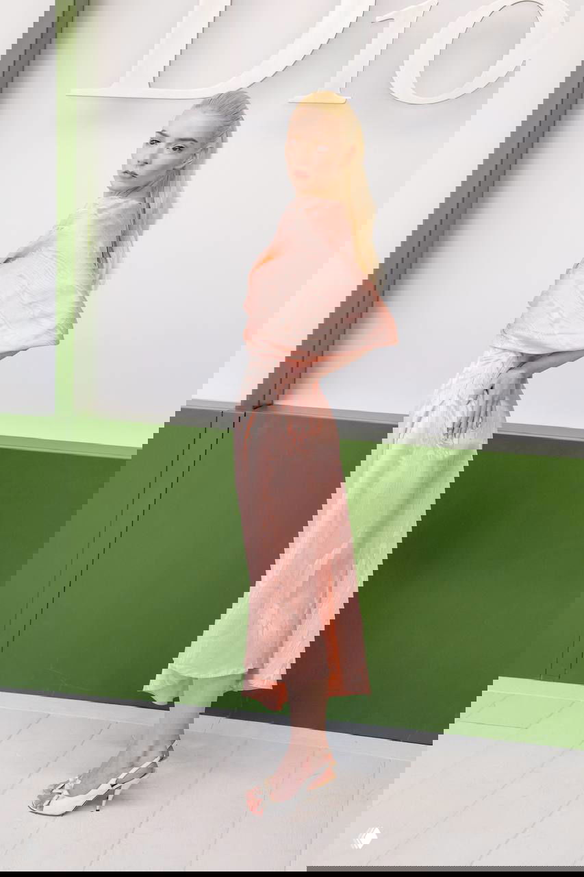 Модель в светло‑персиковом шелковом платье с драпировкой позирует в профиль на фоне белого баннера с крупной надписью бренда.