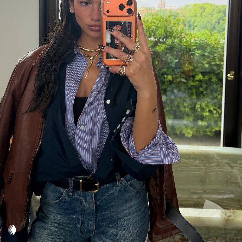 Крупный план селфи Дуа Липы с украшениями на пальцах, полосатая рубашка, кожаная куртка и джинсы — акцент на аксессуары и повседневный стиль.