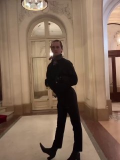 Вертикальный кадр из видео в интерьере здания: мужчина в элегантном костюме позирует в холле с колоннами и мраморной отделкой.