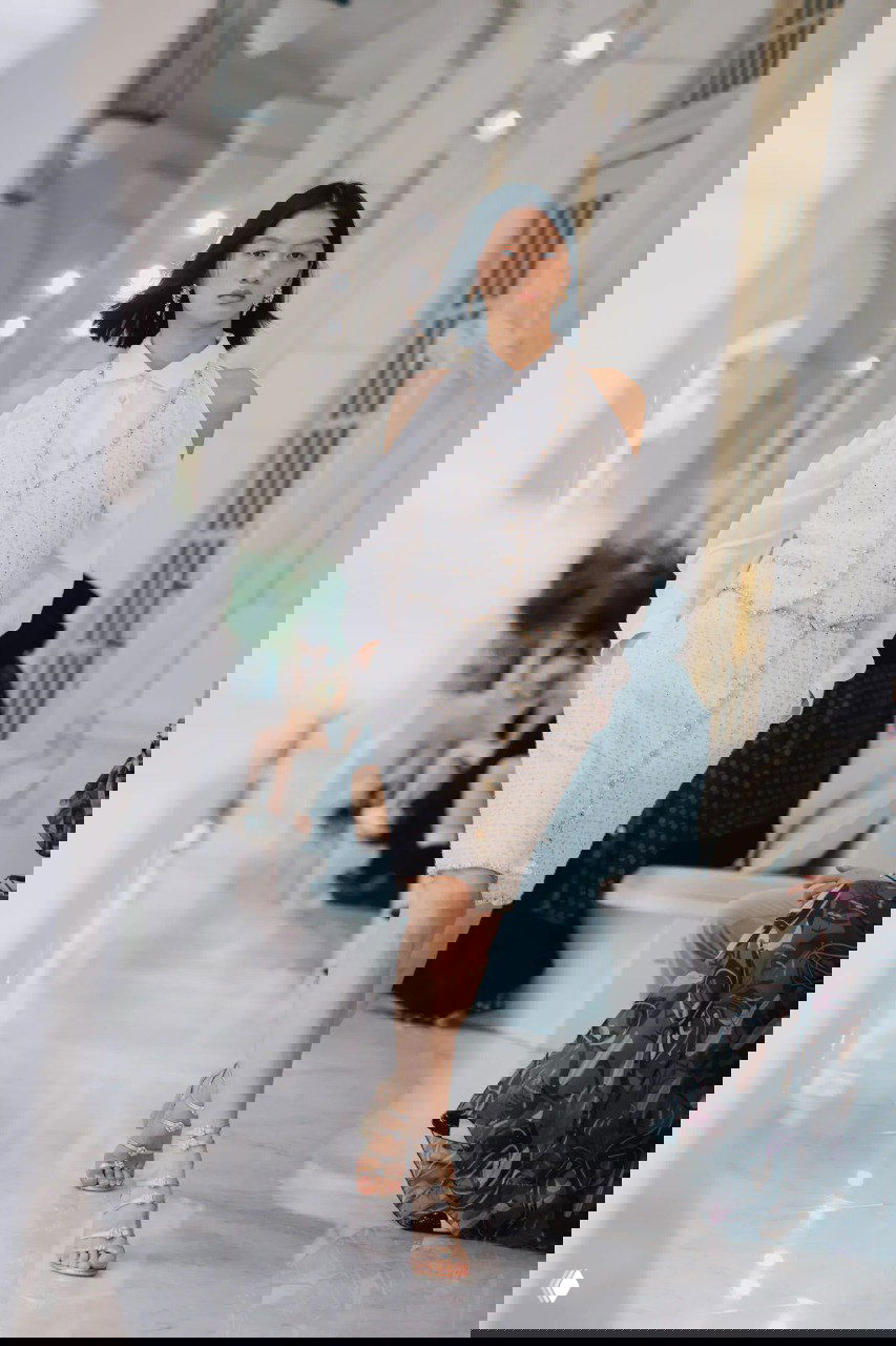 Белый костюм‑жилет и юбка Chanel в пастельной гамме, модель идёт по коридору отеля с колоннами и зрителями во время показа в Сингапуре.