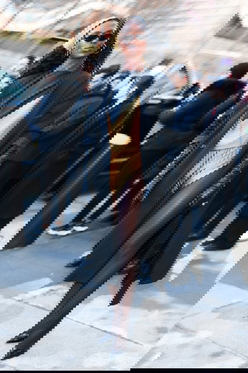 Высокая модель в чёрном длинном пальто, золотистом комбинезоне и длинных перчатках позирует перед камерой возле входа на показ.