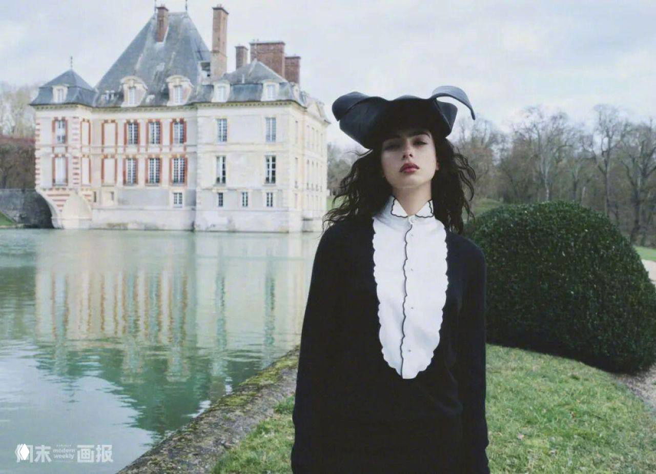 Модель в тёмной одежде и шляпе позирует у пруда на фоне загородного поместья и ухоженного парка в стиле коттеджкор.