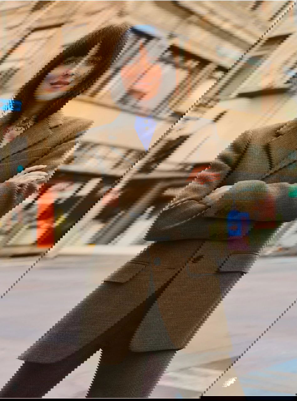 Актриса в твидовом пиджаке и брючном костюме на городском фоне; сжатые руки и уверенная поза подчёркивают классическую эстетику образа.