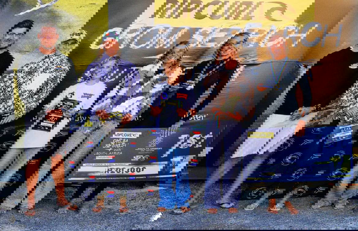 Церемония награждения Rip Curl Grom Search в ЮАР: группа участниц на подиуме с призами и баннером, среди них юная российская серферка Таисия Беляева.