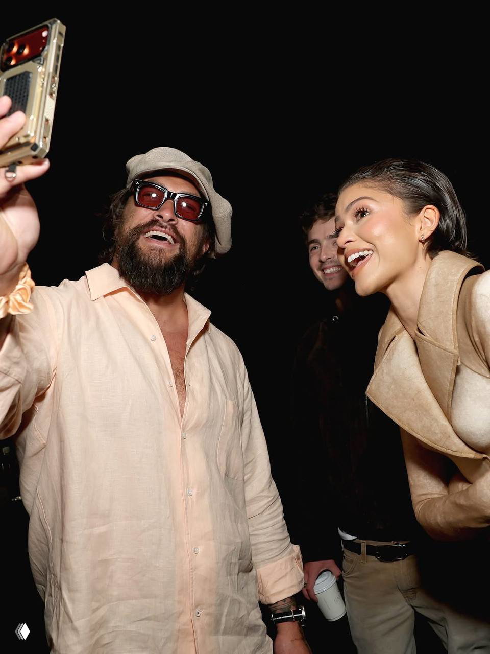 Живой кадр за кулисами: человек с бородой делает селфи на телефон, рядом улыбаются участники презентации, атмосфера неформального общения на мероприятии.