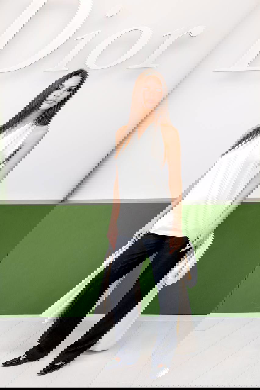 Гостья в белом топе и чёрных широких брюках позирует на бело‑зеленом фоне с крупным логотипом модного дома.