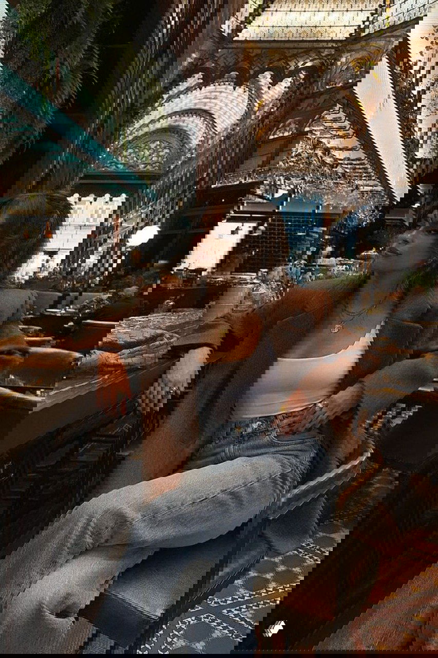 Интерьерное фото женщины в коричневом пальто и джинсах, сидящей у барной стойки — тёплый ресторанный антураж и акцент на аксессуарах