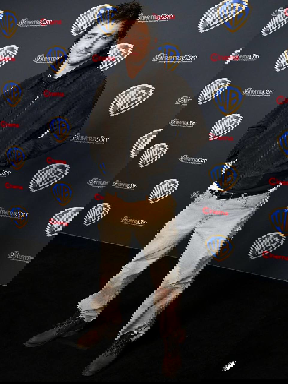 Мужчина в коричневой куртке и поношенных джинсах позирует у пресс-волла Warner Bros, повседневный образ в контрасте с официальной обстановкой CinemaCon.