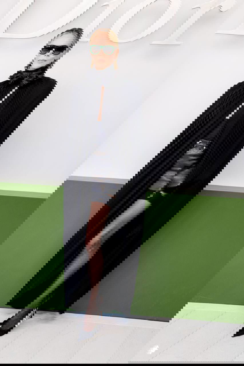 Женщина в длинном чёрном плаще‑накидке и солнцезащитных очках стоит у фотозоны, подчёркивая драматичный вечерний образ.