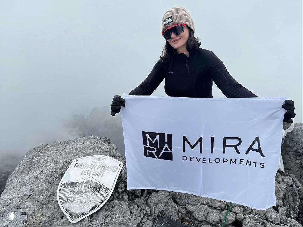 Алина Пекова на вершине держит флаг MIRA Developments и позирует рядом с памятной табличкой: туманная горная сцена и элементы альпинистского снаряжения.