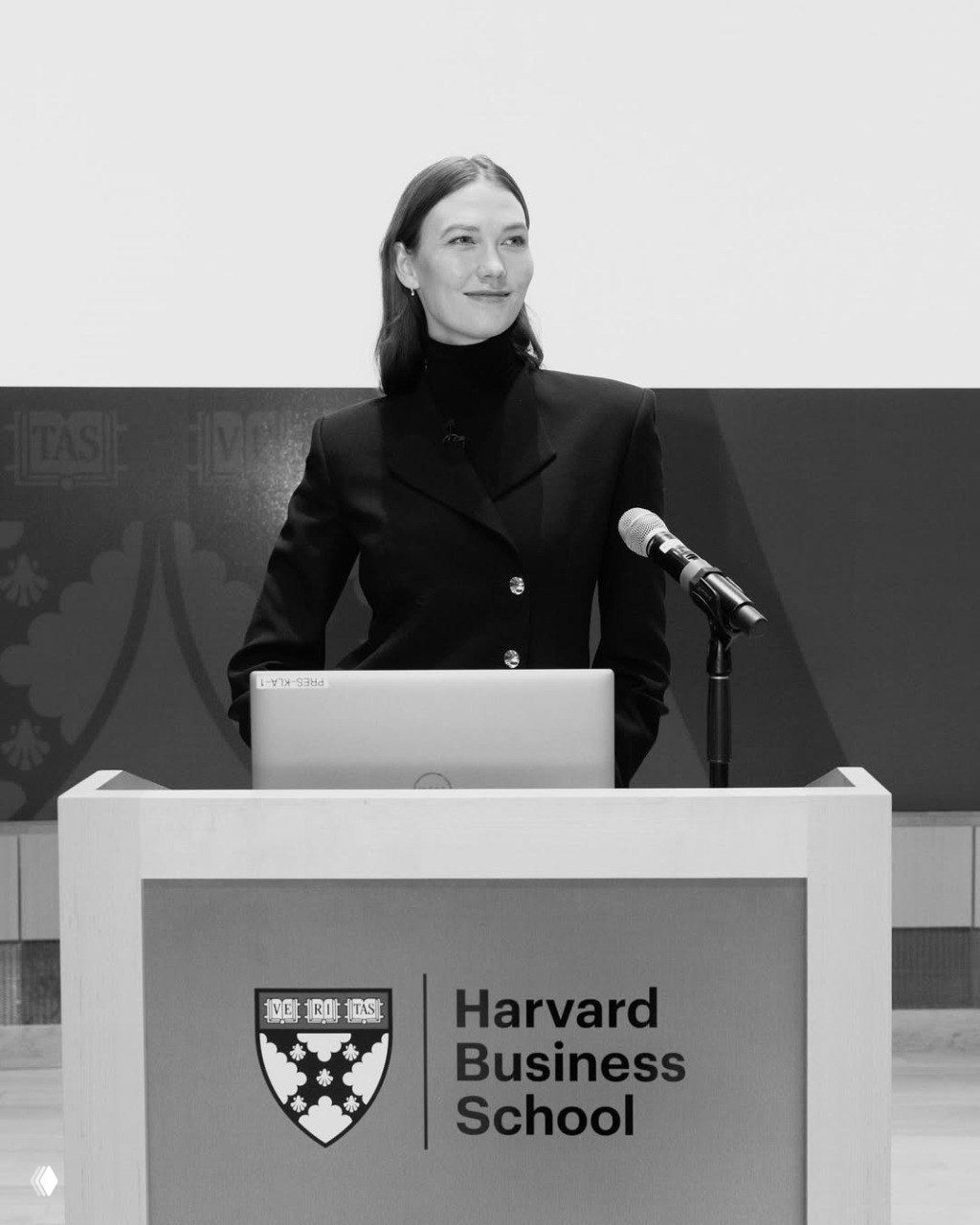 Карли Клосс стоит за кафедрой с логотипом Harvard Business School в деловом образе у микрофона и ноутбука во время выступления на сцене.
