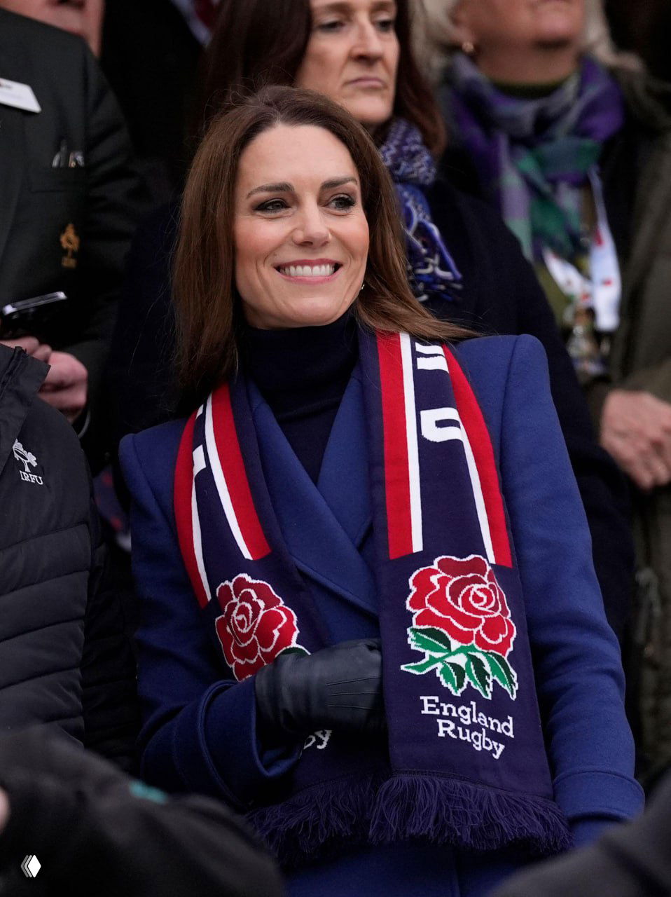 Крупный план женщины в синем пальто и тёмном гольфе с фанатским шарфом England Rugby и перчатками, она улыбается на трибуне.
