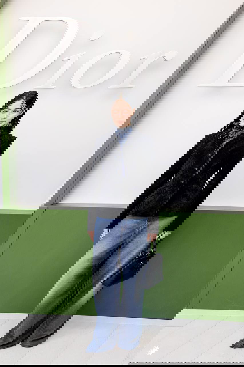 Гостья в чёрном бархатном жакете и широких джинсах с сумкой на фоне логотипа — спокойный элегантный образ для шоу.