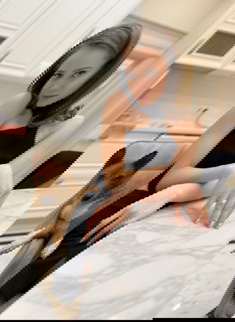 Женщина в элегантном чёрном платье сидит за мраморным столом, рядом на стуле — рыжий пес; кадр похож на светский портрет с питомцем.