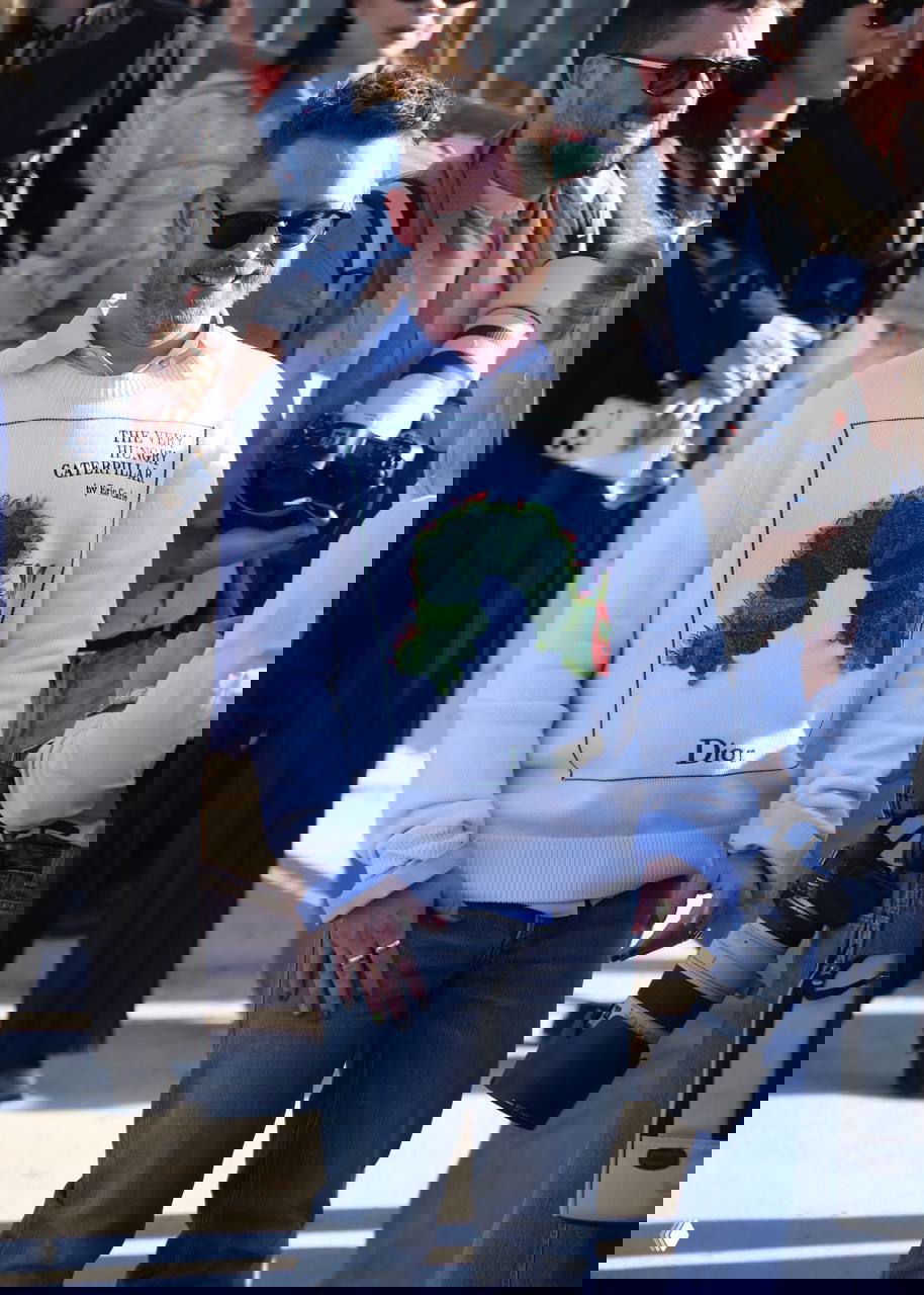 Мужчина в светлом свитере с крупным рисунком и солнцезащитных очках идет по улице возле зоны показа, вокруг фотографы с объективами.