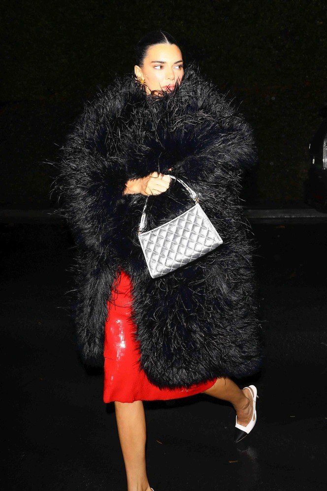 Женщина в большой чёрной пушистой шубе и алом платье идёт по мокрой дороге ночью; серебристая сумка контрастирует с тёмным фоном и блеском платья.