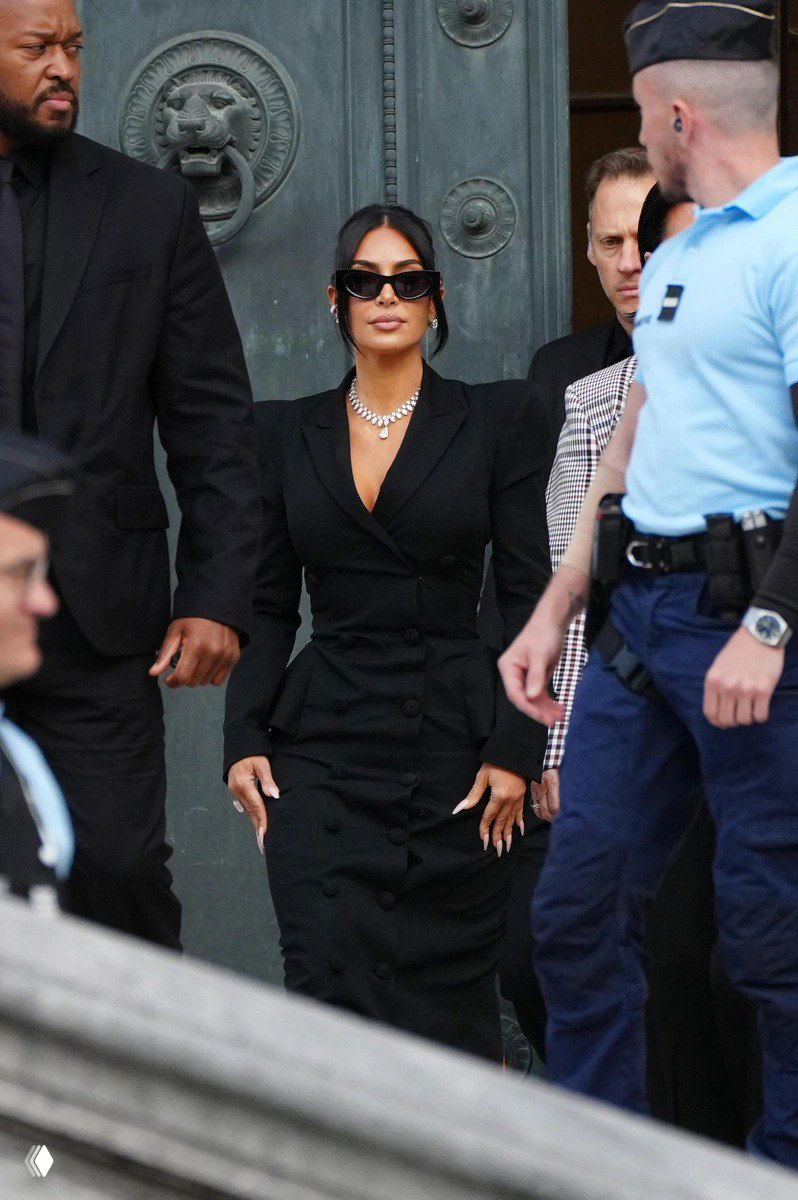 Ким Кардашьян в строгом черном пальто‑платье и солнцезащитных очках среди охраны — образ с деловым акцентом на фасон и силуэт.