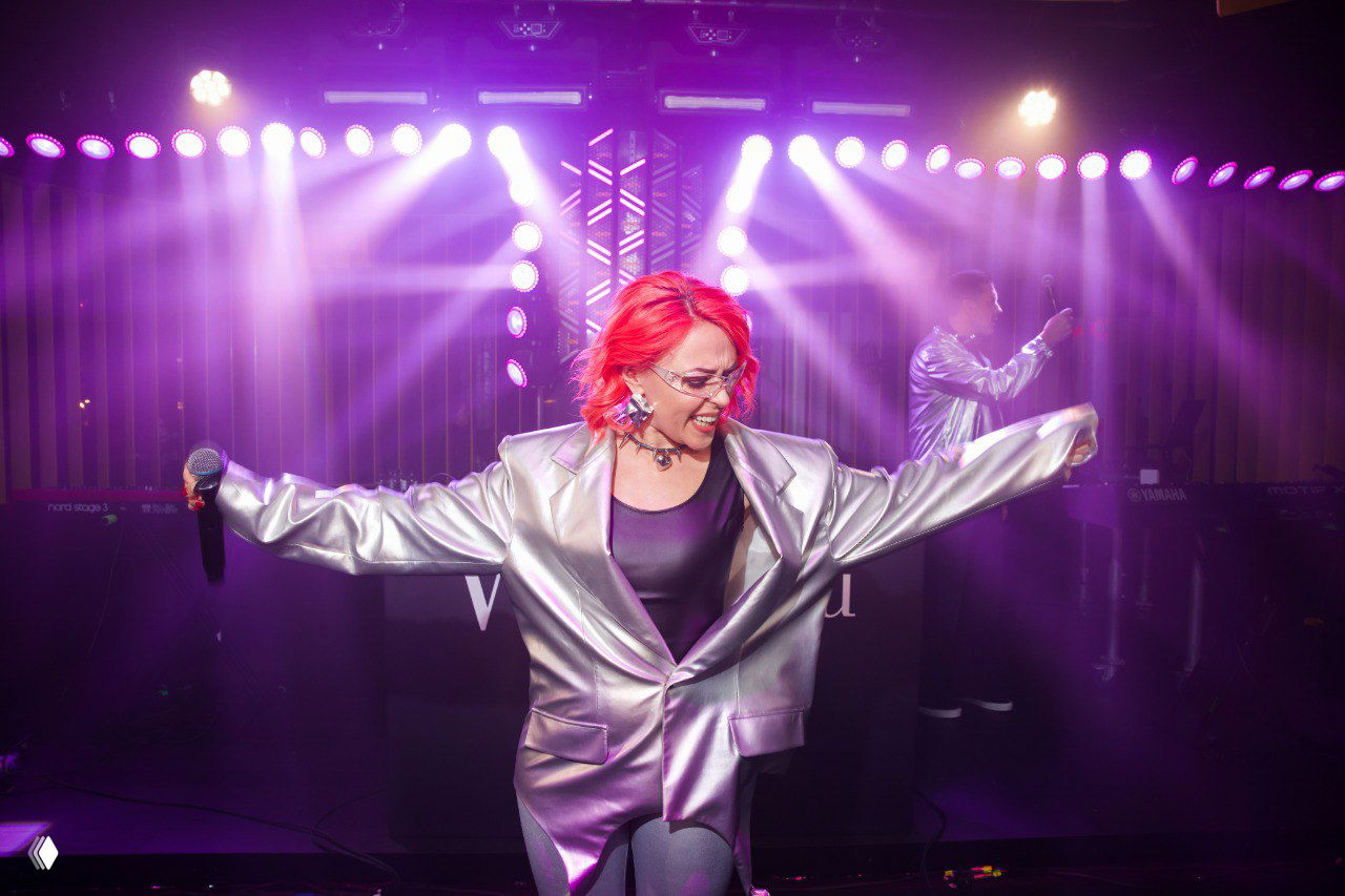 Исполнительница в серебристом пиджаке поёт на сцене под интенсивной фиолетовой подсветкой и эффектами, динамичное выступление.