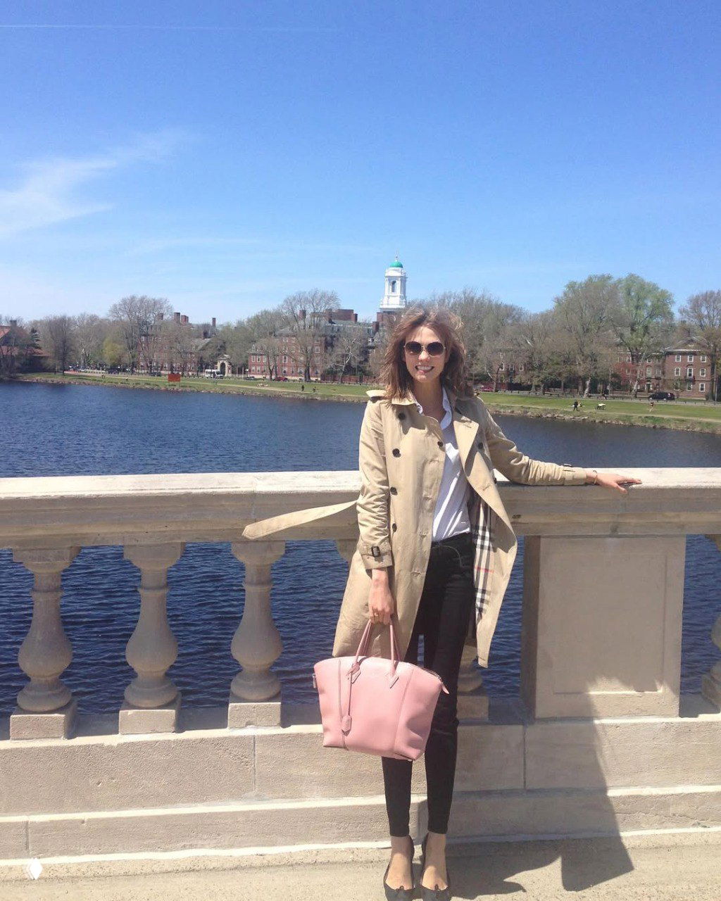 Карли Клосс на прогулке по кампусу Гарварда у реки в лёгком пальто и с розовой сумкой на фоне строений и весеннего пейзажа.