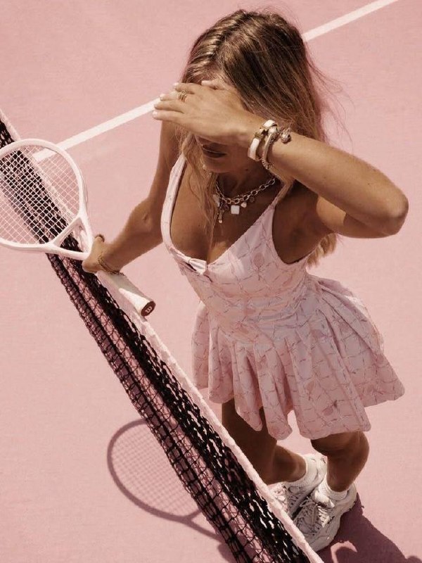 Женщина в розовом теннисном платье у сетки на розовом корте, держит ракетку и прикрывает лицо рукой; акцент на движении и стиле.