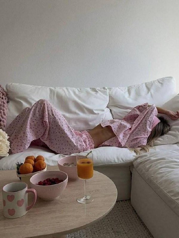 Женщина в розовой пижаме лежит на белом диване рядом с кофейной кружкой, мисками с ягодами и бокалом апельсинового сока — спокойный утренний завтрак