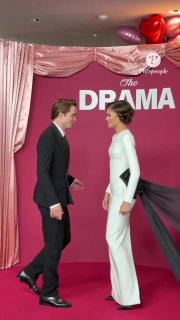 Вертикальный кадр: пара идёт по красной дорожке мимо баннера с надписью премьеры, снимок в мобильном формате подчёркивает атмосферу события в Париже.