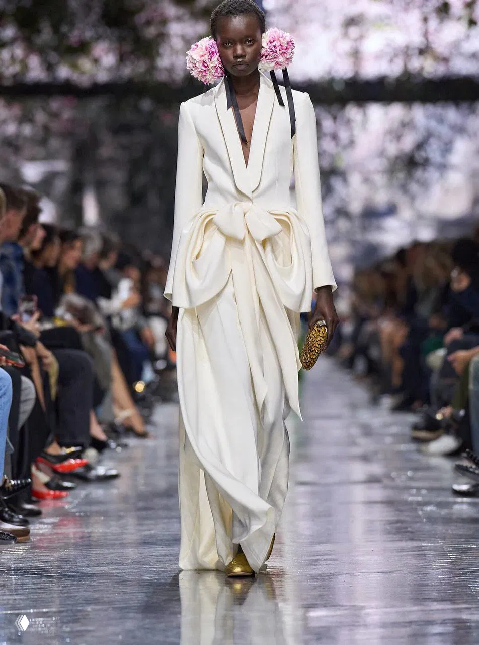 Белое платье с перьями и сложным драпированием; модель на показе первой кутюрной коллекции Джонатана Андерсона для Dior, цветочный фон подиума.