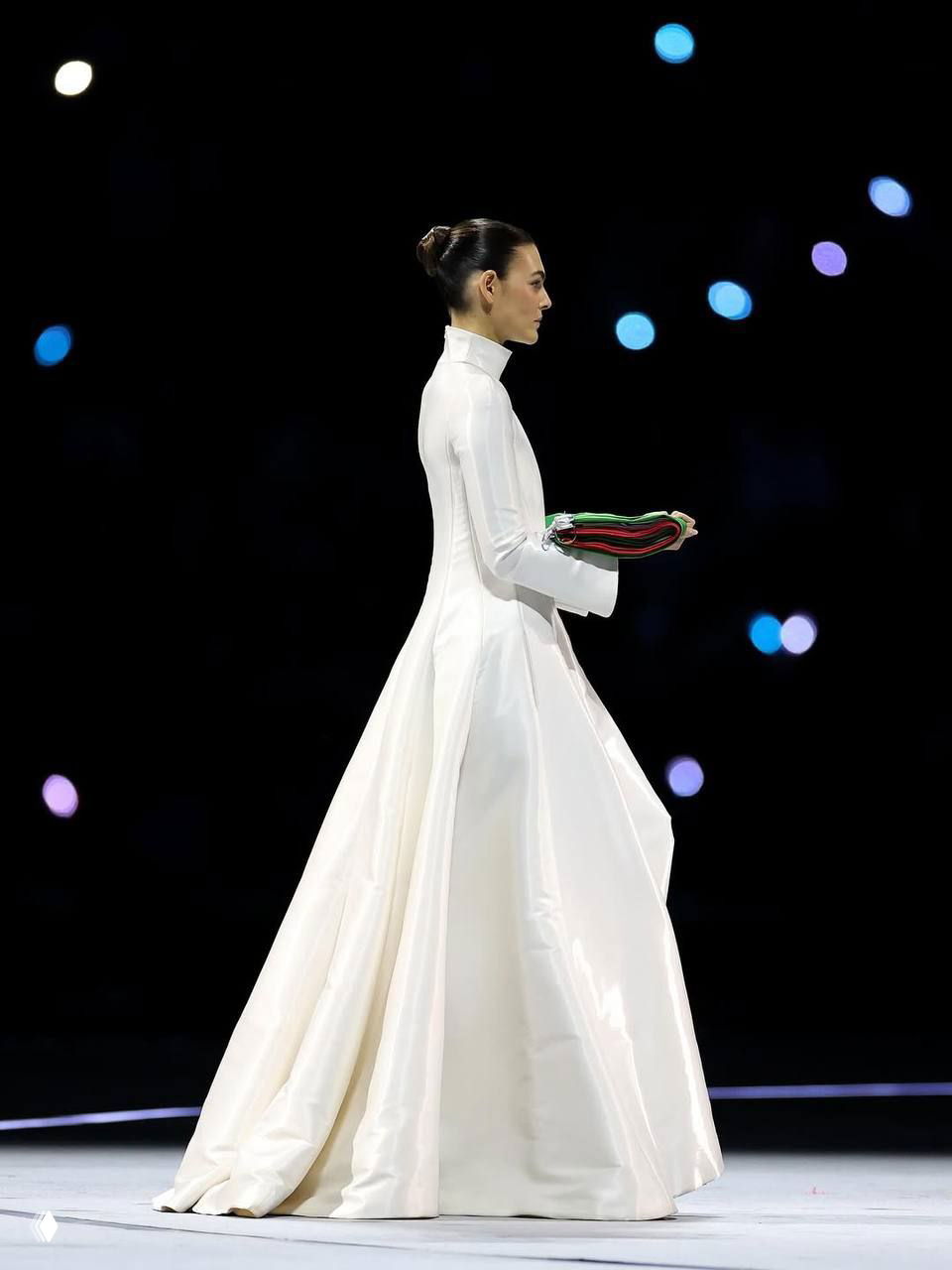 Боковой портрет Виттории Черетти в длинном белом платье от Giorgio Armani, она держит сложенный флаг на тёмном фоне сцены