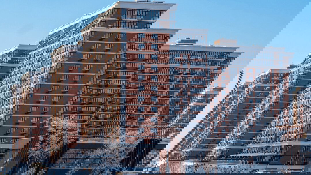 Общий план секций жилого комплекса «Самолет» с панорамным остеклением и балконами; вид на улицу с припаркованными автомобилями и заснеженными деревьями.