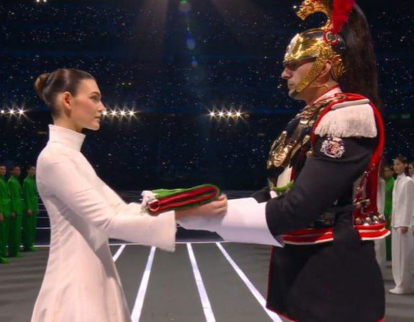 Момент передачи флага: Виттория Черетти принимает символ с руки в парадной форме на фоне олимпийской арены и разметки сцены
