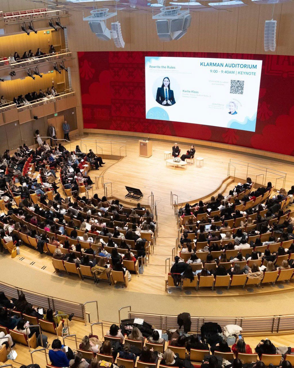 Вид на зал Klarman Auditorium в Harvard Business School: полный зал слушателей, круглая сцена и большой экран с афишей выступления Карли Клосс.