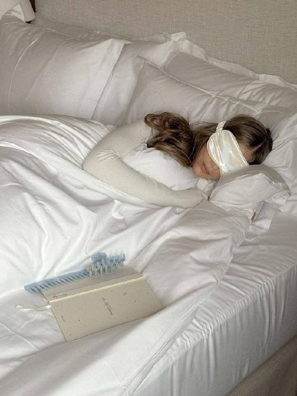 Женщина спит с шелковой маской на лице на белой кровати рядом с дневником и расческой — сцена спокойного сна и утренних ритуалов перед пробуждением