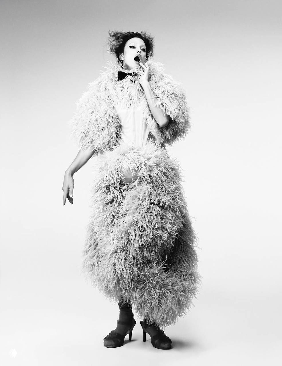 Чёрно‑белый снимок: Ирина Шейк в плотном пёрышном платье, театральная поза и движение ткани, эстетика высокой моды в студии.