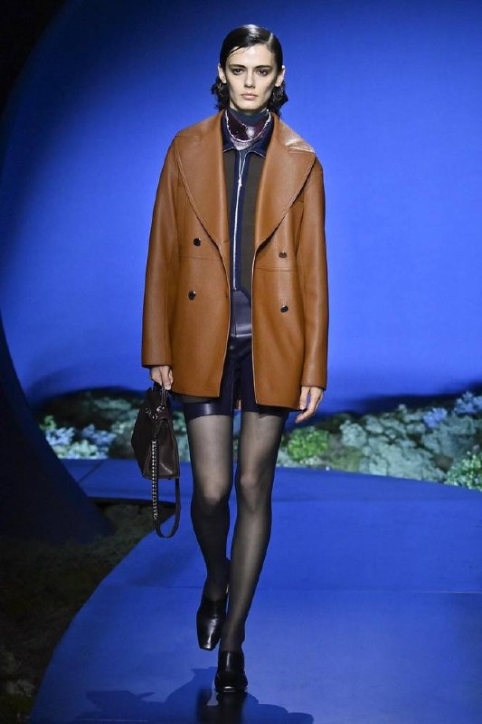 Модель в табачном кожаном пальто Hermès и мини‑образе несёт сумку тёмного оттенка на фоне синего «лунного» декора подиума, сочетая кожу и замшу.