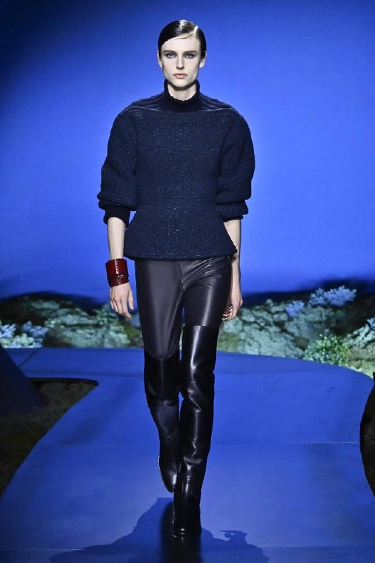 Модель в тёмно‑синем вязаном свитере и кожаных брюках Hermès идёт по подиуму, подчёркивая игру фактур и глубокую сумеречную палитру коллекции.