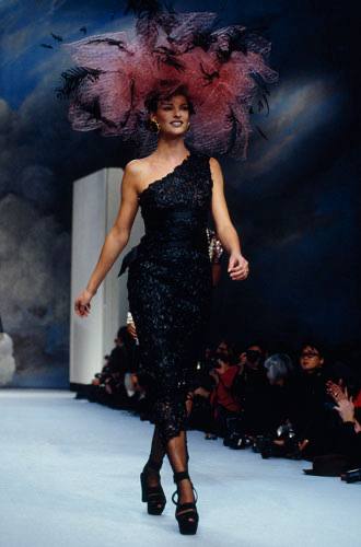 Подиум: модель в чёрном платье и крупной декоративной шляпе, драматический образ показа мод с затемнённой публикой.