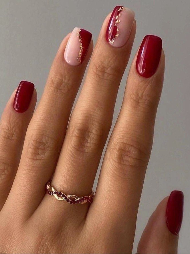 Рука крупным планом с комбинацией глубокого красного покрытия и нюдовых акцентных ногтей, украшенных тонкой золотой крошкой.