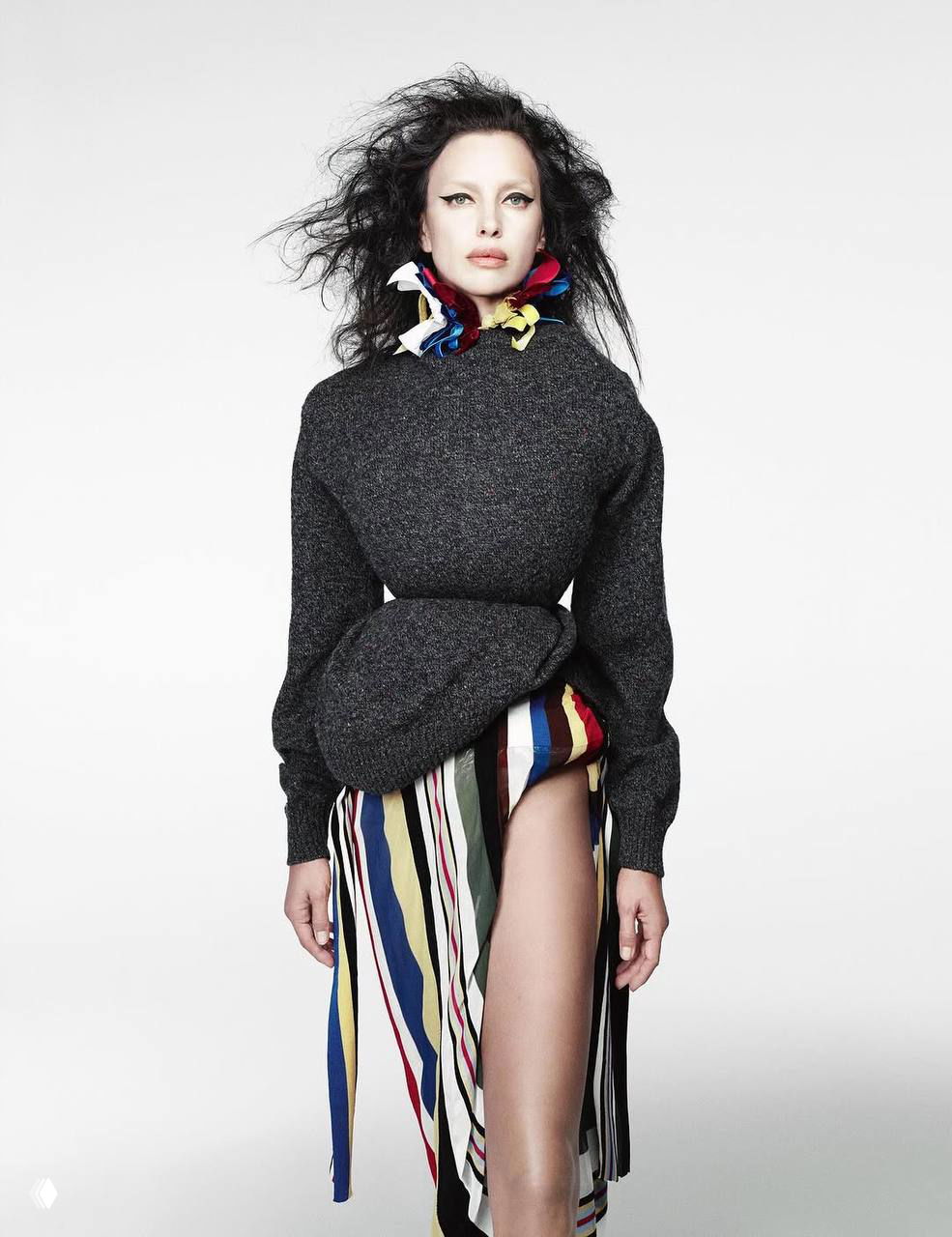 Ирина Шейк в асимметричной юбке с цветными лентами и тёмном свитере, стройная модельная поза в полный рост на нейтральном светлом фоне.