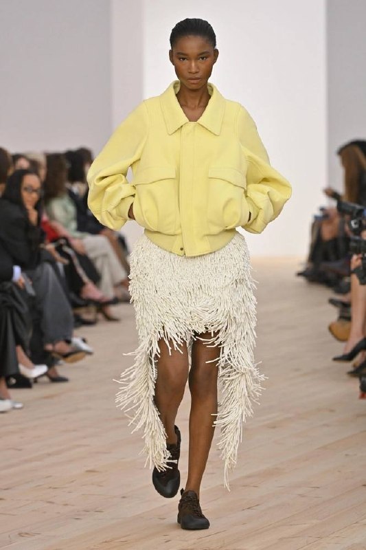 Модель в кремовой юбке с декоративной бахромой и светло-жёлтой куртке‑бомбере, образ в стиле casual на подиуме с публикой по бокам.