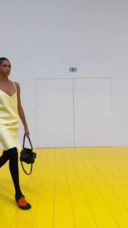 Компактный кадр: модель в жёлтом лёгком платье и контрастных оранжевых туфлях идёт по зеркально‑жёлтому полу подиума, минимализм сцены подчёркивает образ.