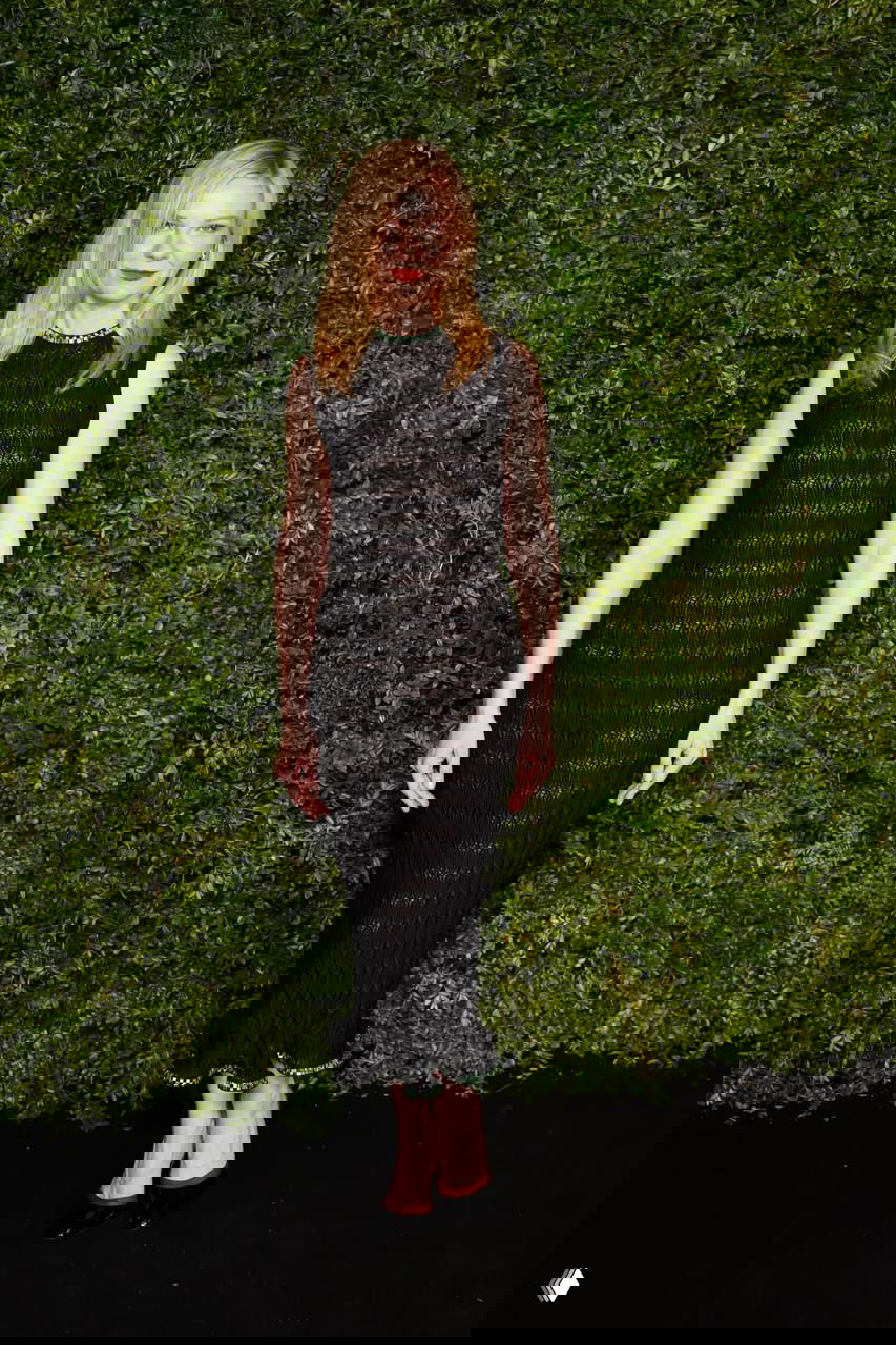 Женщина в приталенном чёрном платье и украшении на шее позирует на фоне зелёной растительной стены во время вечеринки Chanel.