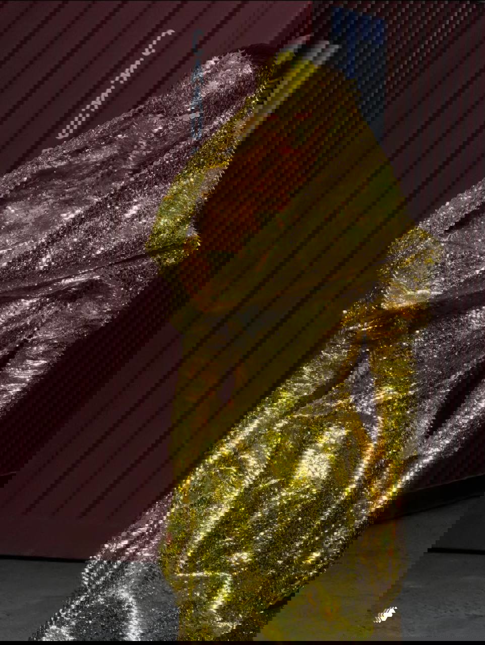 Модель в золотом платье и маске Maison Margiela: жатый металлизированный материал, плотный блеск и театральный образ на фоне контейнеров.