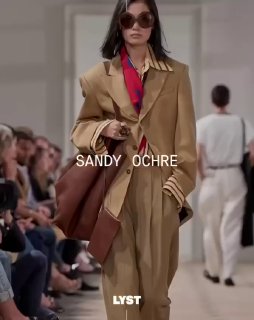 Модель в песочно-охристом свободном костюме идёт по подиуму; на кадре подпись «SANDY OCHRE» и логотип LYST, атмосфера показа и зрители.